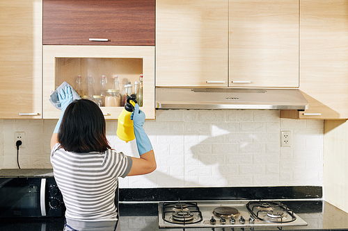 Young woman spraying disinfecting detergent on doors of kitchen cabinets and wiping it with soft cloth, view from the back