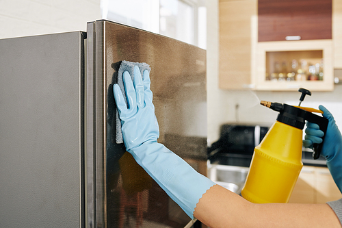 Close-up image of woman in rubber gloves cleaning stainless steel refrigerator with cloth and spray bottle