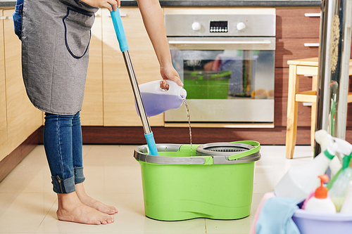 Housewife in apron pouring floor cleaning liquid with lavender scent in bucket with mop