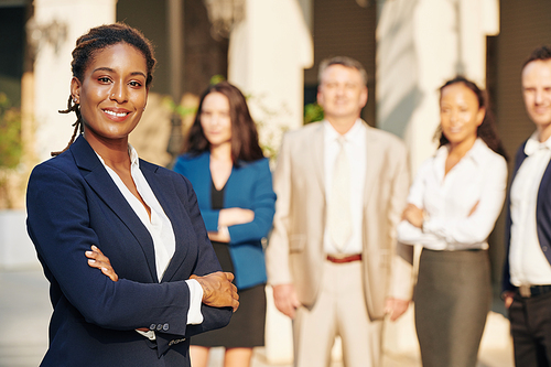 Beautiful confident successful businesswoman standing with arms folded and smiling at camera, her colleagues standing in background
