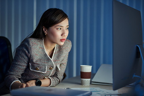Serious young pretty business lady reading document on glowing screen when working in office late at night