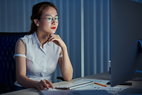 Pretty young Vietnamese businesswoman in glasses working on computer in office late at night