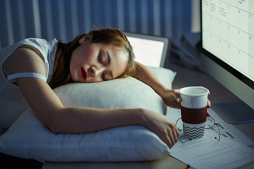 Female project manager sleeping at her office desk with coffee cup in hands and calendar on computer screen