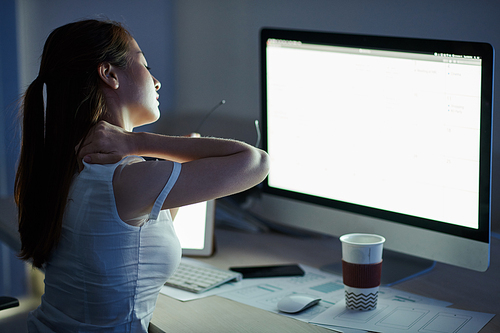 Young businesswoman kneading stiff neck when sitting at glowing computer screen in dark office