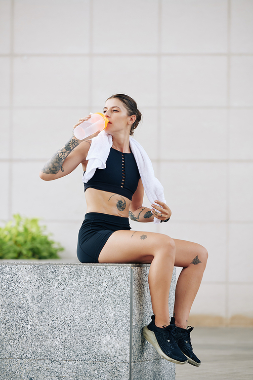 Beautiful fit young woman resting on parapet and drinking fresh water after exhausting outdoor training