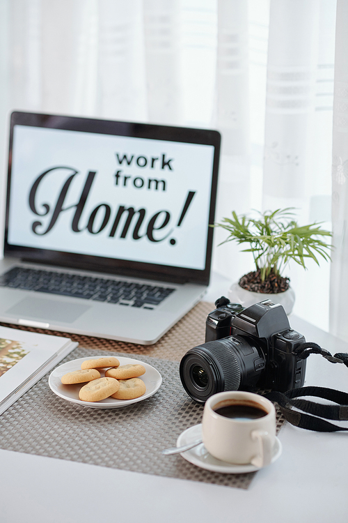 Desk of photographer working from home during coronavirus pandemic with cup of coffee, plate with cookies and laptop