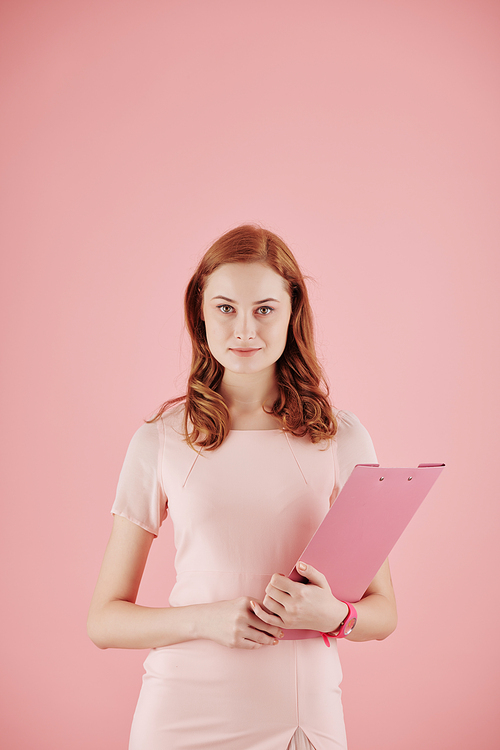 Portrait of pretty young red haired businesswoman with folder posing against pink background