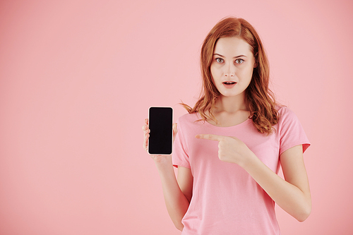 Portrait of shocked young woman with red hair pointing at smartphone screen