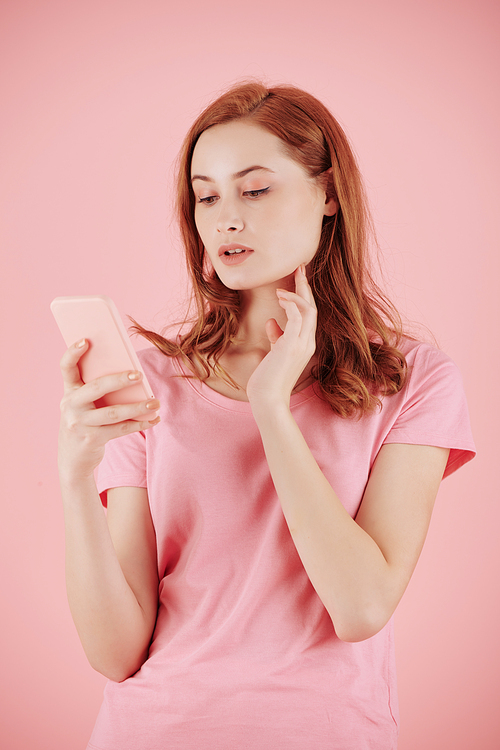 Beautiful young red haired woman reading text messages on smartphone, isolated on pink