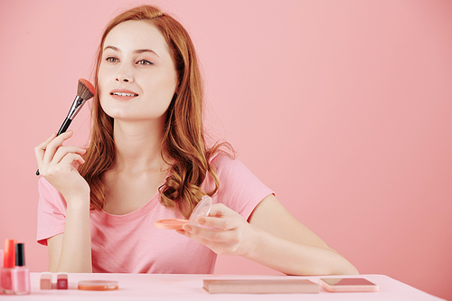 Beautiful smiling young woman applying pressed blush when sitting at vanity and doing makeup