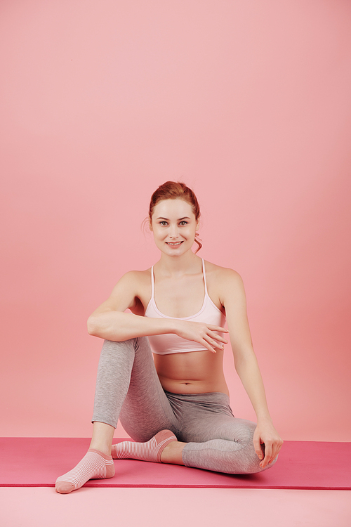 Pretty smiling young woman resting on yoga mat after exercising in pink studio