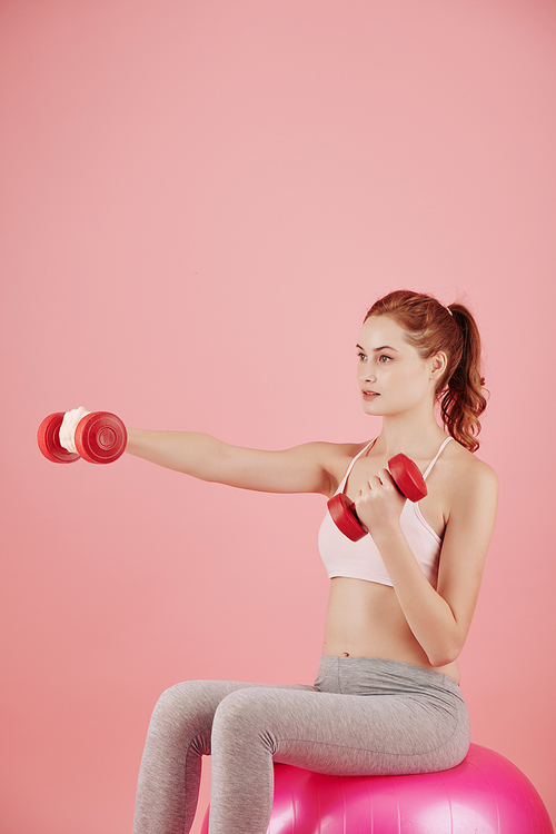 Serious pretty young woman sitting on pink fitness ball and doing exercise with dumbbells