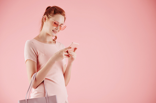 Attractive smiling young woman in sunglasses texting friends, isolated on light pink