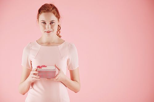 Portrait of smiling young pretty red haired woman with giftbox standing against pink background