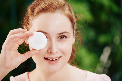 Face of smiling young woman covering eye with cotton pad and looking at camera