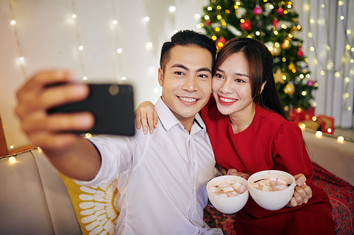 Smiling beautiful Vietnamese couple taking selfie with cup of hot chocolate against Christmas tree at home