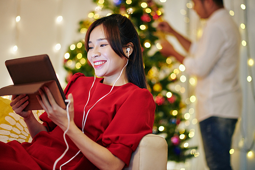 Happy pretty young Vietnamese woman resting on sofa and video calling her friends when husband decorating Christmas tree in background