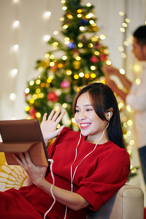 Beautiful young Vietnamese woman video calling her friends when boyfriend decorating Christmas tree in background