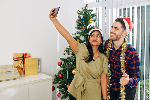 Cheerful multi-ethnic colleagues in Santa Claus hats taking selfie at Christmas tree in office