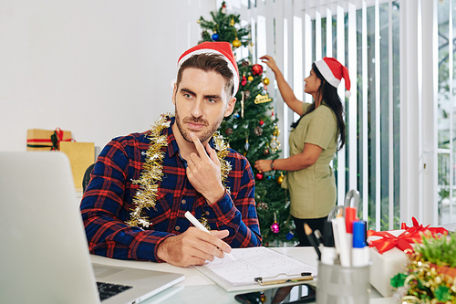 Pensive young businessman in Santa hat working on annual report when his colleague decorating Christmas tree in office