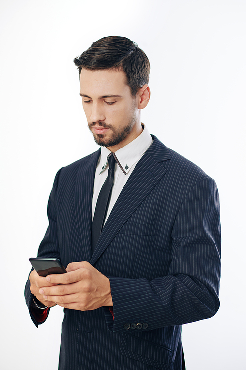 Successful serious young entrepreneur in suit reading text messages from coworkers on smartphone