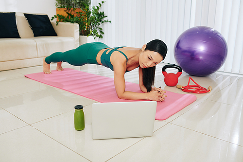 Slim Asian woman standing in plank position when following fitness tutorial on laptop screen