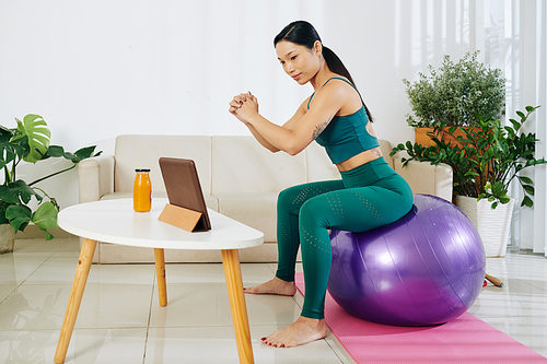 Young woman training on fitness ball at home when watching tutorial on digital tablet