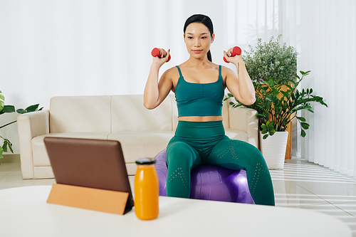 Fit pretty young Asian woman sitting on fitness ball, watching video guide on tablet and doing exercise with dumbbells