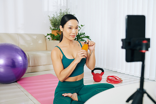 Pretty smiling Asian fitness blogger recording juice commercial for her channel at home