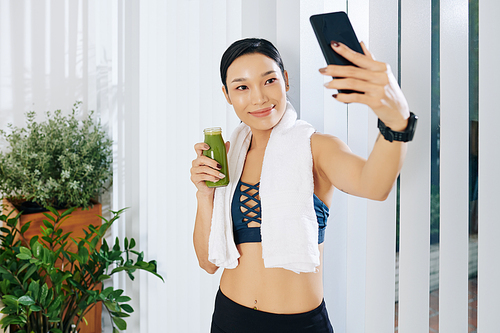Pretty smiling young Asian woman drinking green smoothie after training at home in the morning an taking selfie
