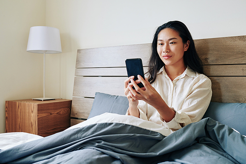 Smiling young Chinese woman sitting in bed after waking up and reading text messages in her smartphone