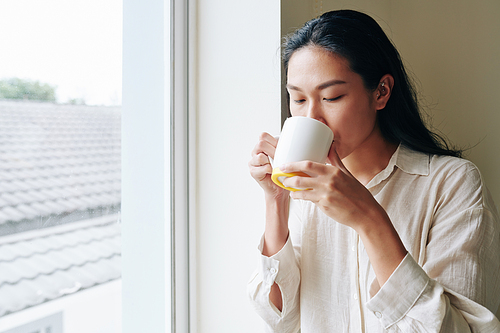 Beautiful young Chiense woman enjoying big mug of morning coffee when standing at window