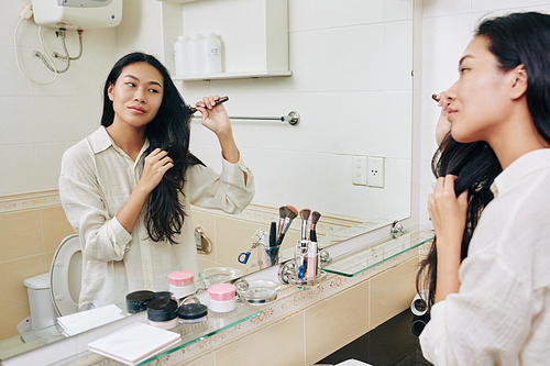Smiling pretty young Chinese woman brushing her long hair in front of bathroom mirror