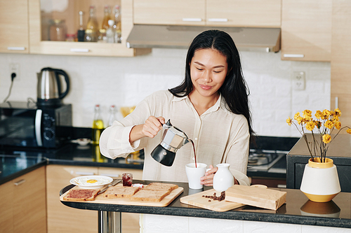 Smiling pretty Chinese woman pouring coffee in cup when eating breakfast in kitchen