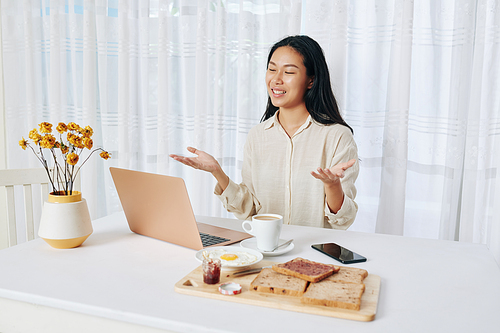 Pretty young businesswoman sitting at her table with breakfast and having online meeting with colleague