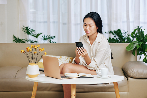 Pretty young Asian businesswoman in pajamas sitting of sofa after working on laptop and checking messages from colleague in smartphone