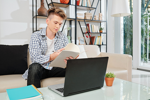 Student checking notes in his textbook when doing homework on laptop at home