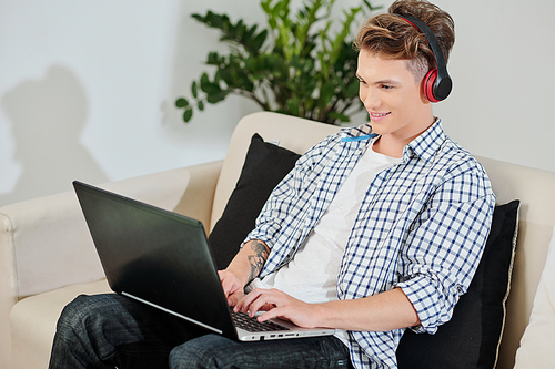 Smiling teenage boy in headphones sitting on sofa at home and coding on laptop