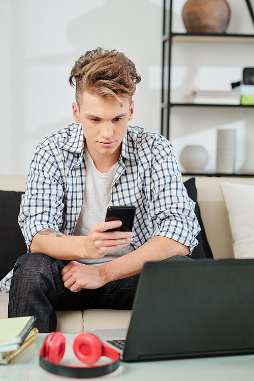 Serious teenager sitting on sofa at home, reading text messages or posts on social media on his smartphone