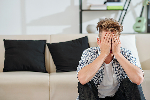 Crying frustrated teenage boy sitting on the floor in living room and covering face with hands