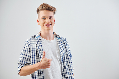 Portrait of joyful teenage boy with toothy smile showing thumbs-up and looking at camera