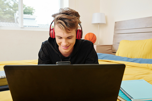 Smiling teenage boy sitting on bed with opened laptop and checking text messages on his smartphone