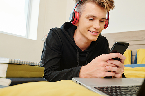 Smiling teenage boy listening to music in headphones and playing game on smartphone when resting in his room