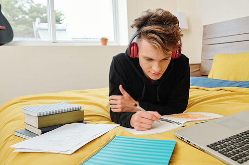 High school student lying on bed at home and writing in copybook when doing homework for math class