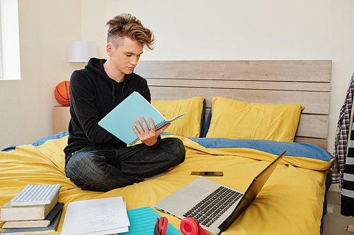 Software engineering student sitting on bed with books, laptop and textbooks around and writing in notebook