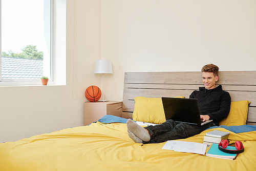 Smiling teenage boy in hoodie sitting on bed with laptop, coding, playing game or attending online class