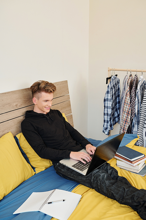 Smiling teenage boy sitting on bed in college dormitory and working on laptop with books and textbooks around