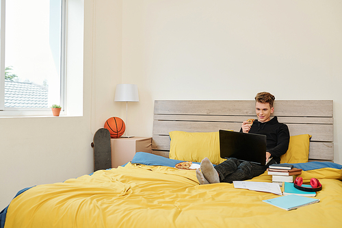 Positive high school student sitting on bed, eating pizza and watching online webinar when staying home during quarantine