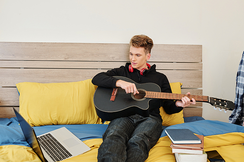 Smart teenage boy watching lesson online on his laptop and learning how to play guitar