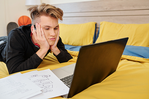 Bored college student lying on bed, watching online class on programming and drawing flowcharts in copybook